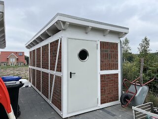 Kleines weißes Gartenhaus mit Fachwerkfassade, Pultdach und runder Türöffnung